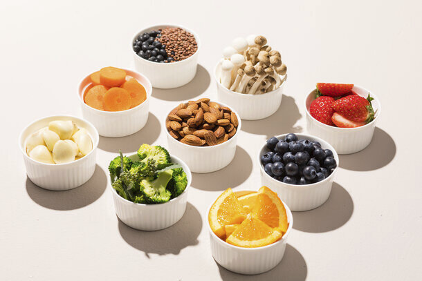 A neatly arranged assortment of small white ramekins filled with various healthy foods, including sliced carrots, blueberries, strawberries, orange wedges, broccoli, almonds, garlic cloves, and mushrooms, placed on a light surface