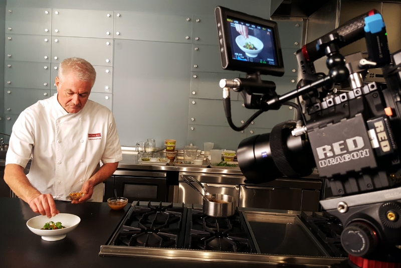 A chef in a professional kitchen is being filmed while preparing a dish, highlighting culinary techniques and ingredients.